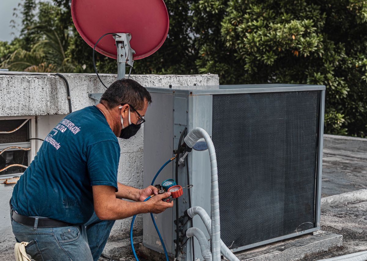 An HVAC technician kneeling on a rooftop servicing an outdoor air-conditioning unit with refrigerant gauges