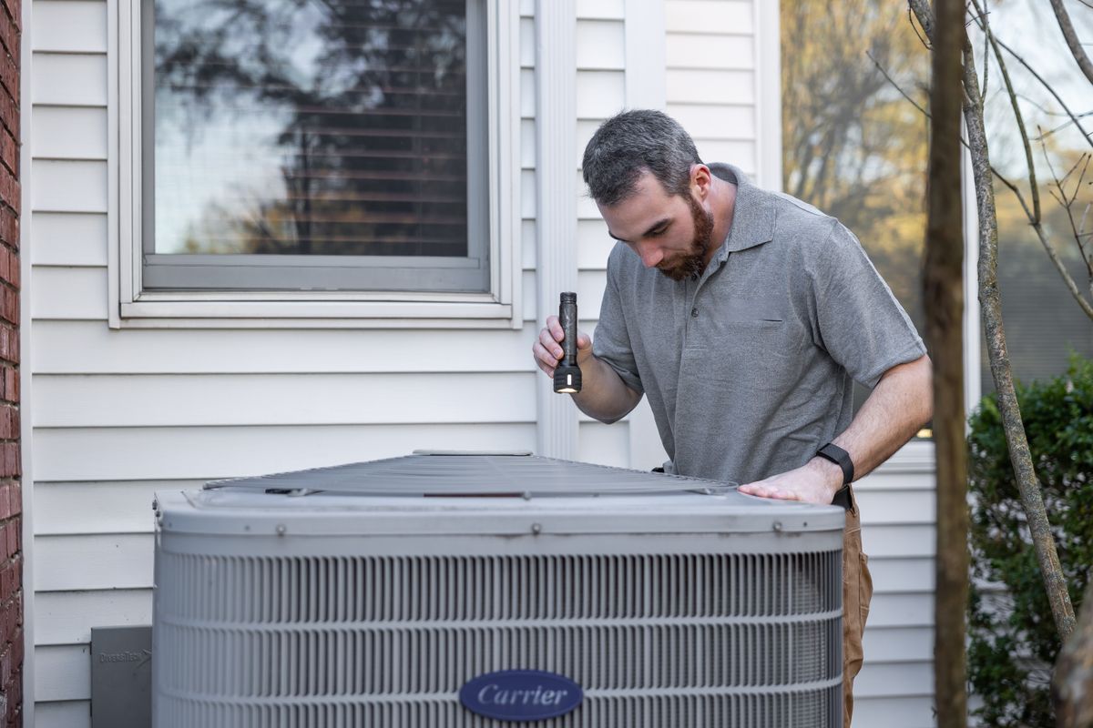 An HVAC technician crouched beside an outdoor air-conditioning condenser unit performing maintenance