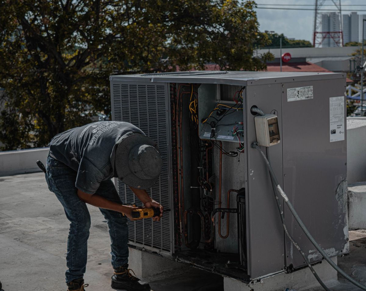 An HVAC technician crouched on a rooftop servicing an outdoor air-conditioning unit with hand tools